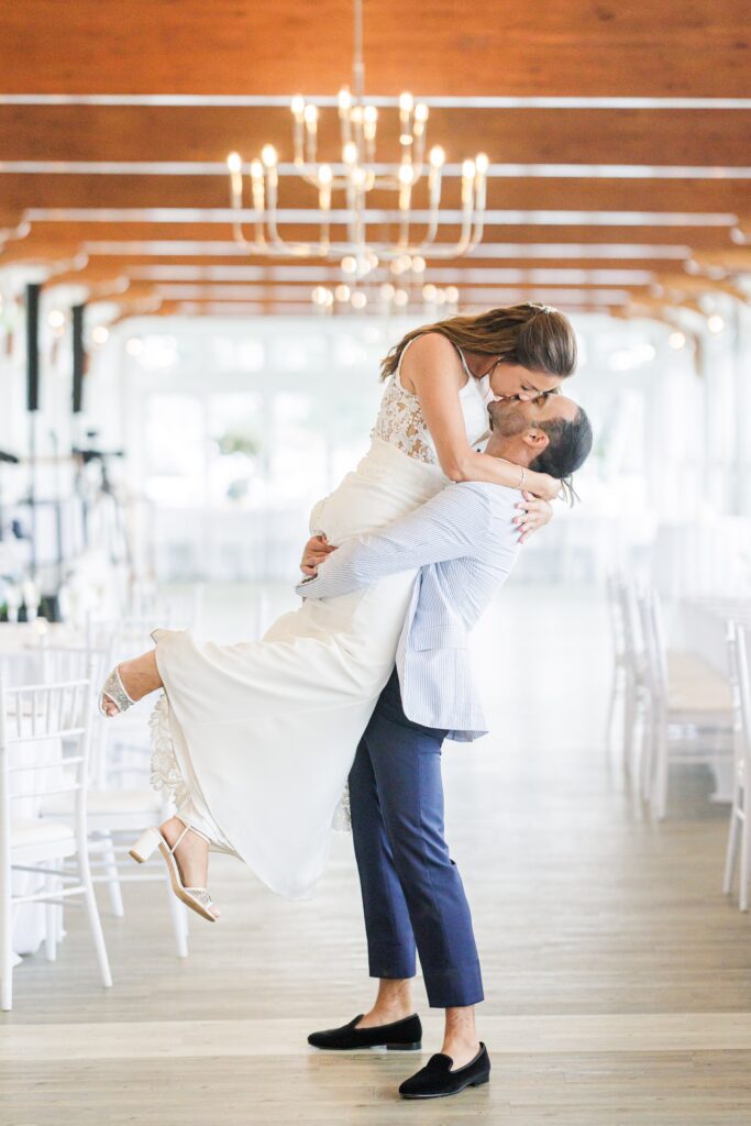 A groom lifts the bride inside a bright, elegant wedding venue with chandeliers and white chairs. The bride leans down to kiss him, smiling as her flowing white dress drapes down. Wychmere wedding photographer