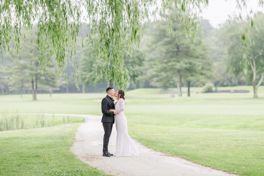 A bride and groom share a kiss on a winding path surrounded by lush green grass and trees. The bride wears a fitted white gown, and the groom wears a black suit with a red boutonnière. Drooping branches frame the top of the image. Cape Club of Sharon wedding