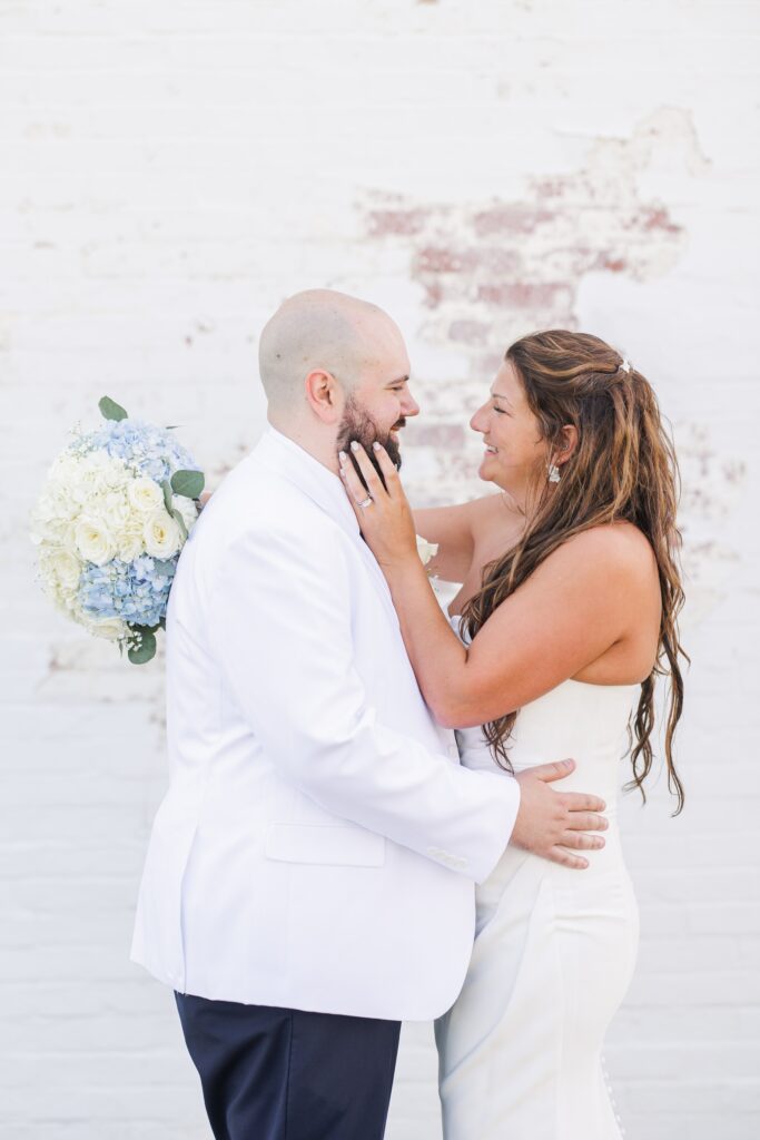 A newlywed couple stands close together in front of a white brick wall. The groom, in a white suit, smiles at the bride as she touches his face affectionately. The bride holds a bouquet of white and blue flowers. Dennis MA Cape Cod wedding photographer