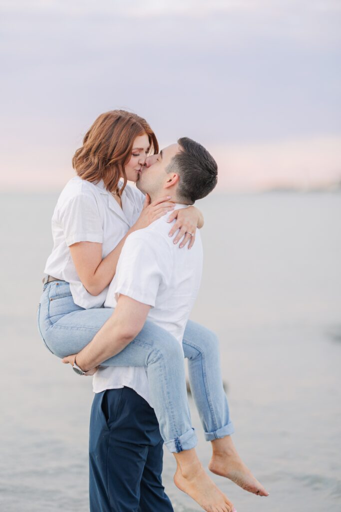 A couple shares a kiss near the water at sunset. The man lifts the woman into his arms as she wraps her arms around his neck. They are both dressed in casual white shirts and jeans, with soft pastel hues in the sky. Wingaersheek Beach