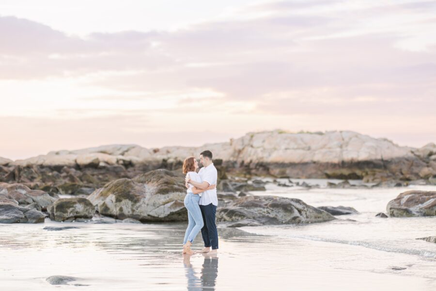 A couple stands on a reflective shoreline at sunset, embracing as gentle waves surround them. The woman wears jeans, and the man wears a white shirt. Rocks and pastel skies fill the background. Wingaersheek Beach