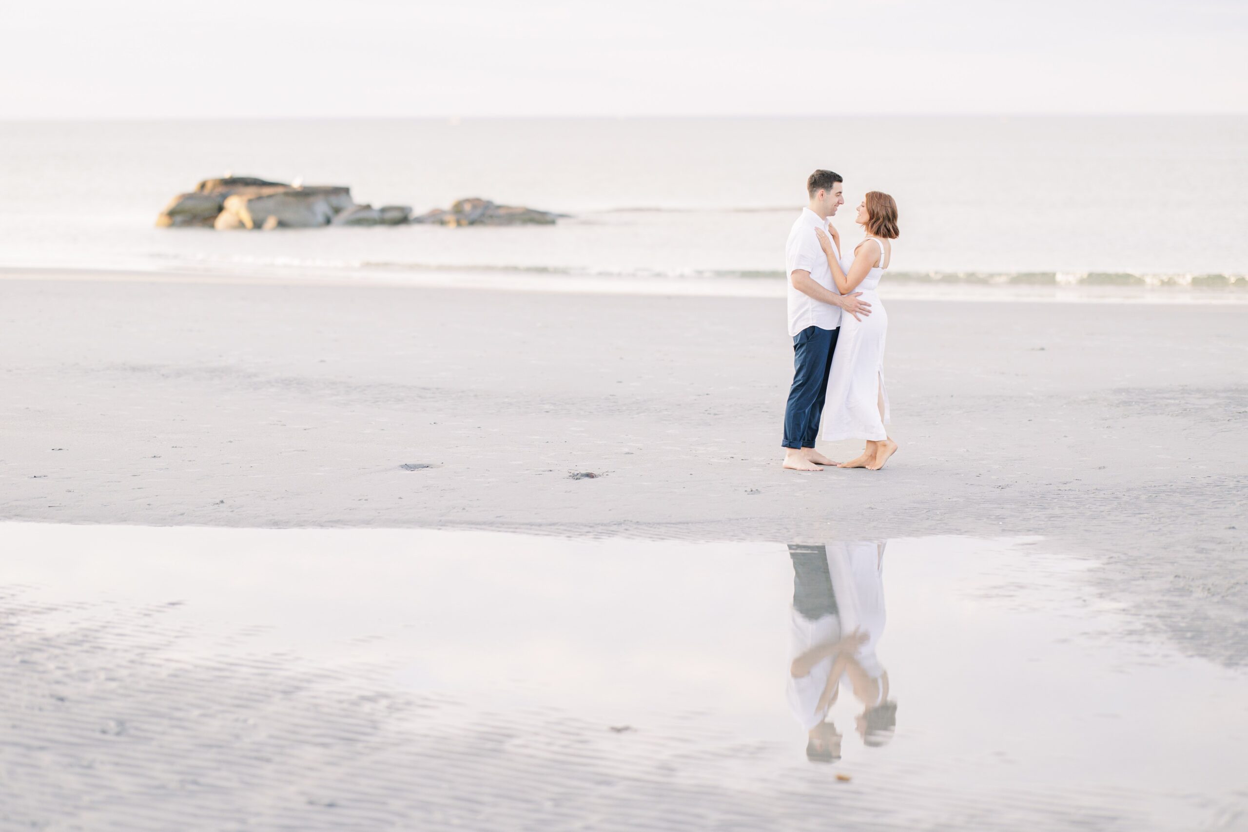 A couple stands barefoot on a quiet beach, embracing each other. Their reflection appears in a still pool of water on the sand. The calm ocean stretches out to the horizon in the background. Wingaersheek Beach