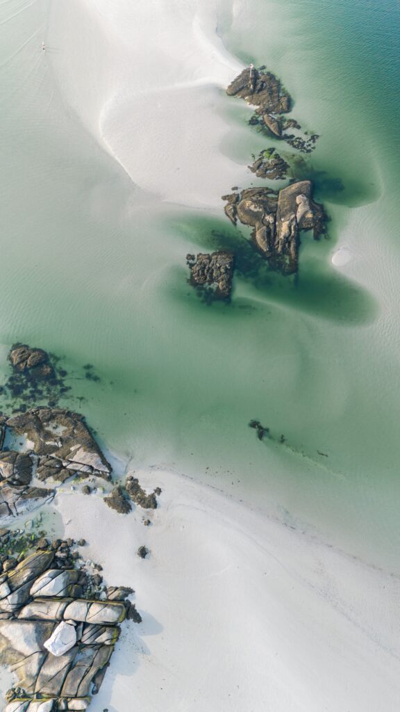  An aerial view of a coastal landscape features clear green water, sandy beaches, and scattered rocky formations. Light and shadow highlight the contours of the rocks and water.