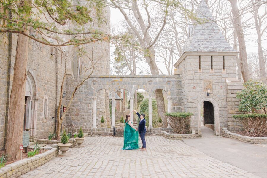 A couple dances in a cobblestone courtyard surrounded by a medieval-style stone castle with archways and a tower. The woman wears a flowing green gown, and the man wears a suit, as they twirl under tall, leafless trees. Hammond castle