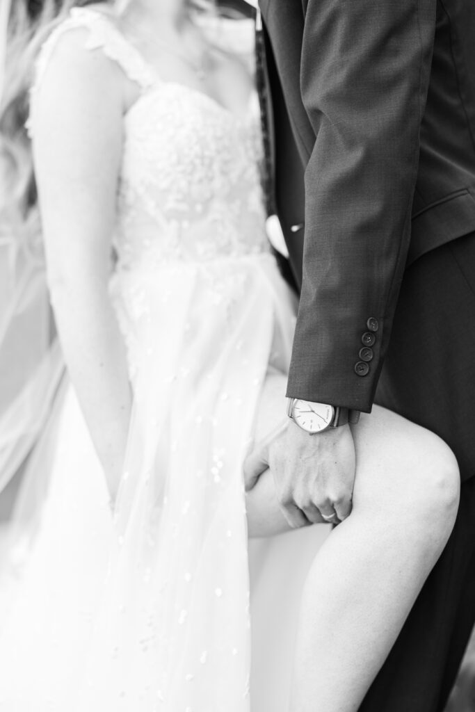 A black-and-white close-up of a groom holding the bride’s leg. The bride’s lace wedding dress is visible, along with the groom’s suit and wristwatch, creating an intimate and artistic composition. Bedford Village Inn wedding photographer