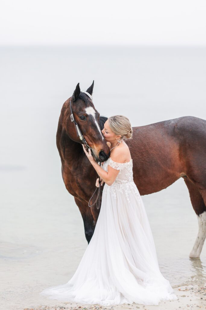 A bride in an off-the-shoulder white wedding gown stands next to a dark brown horse on a calm beach. She kisses the horse’s face while holding its reins, with gentle waves in the background. Cape Cod wedding photographer