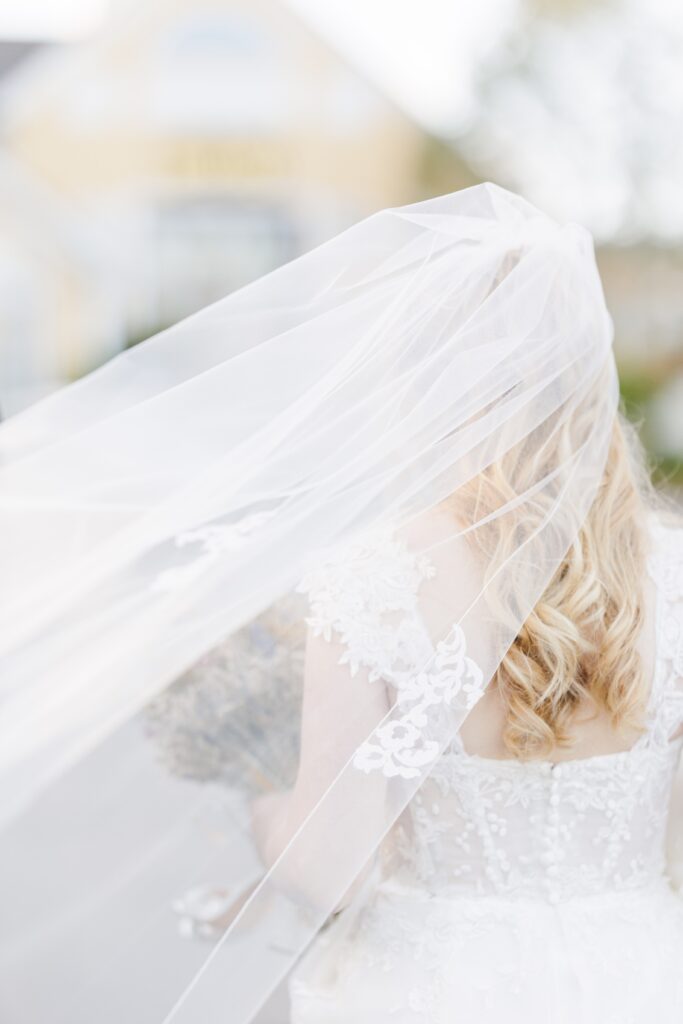 A close-up of a bride’s back as she walks, with her delicate, flowing veil blowing in the wind. Her long blonde hair cascades in soft curls beneath the lace-trimmed veil, and blurred buildings are in the background. Bedford Village Inn wedding photographer