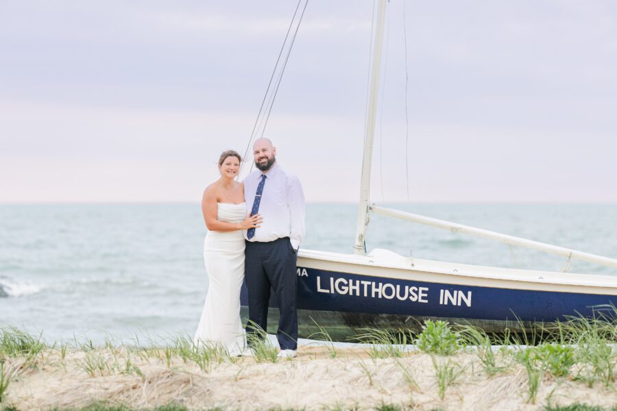 A bride and groom stand together beside a sailboat named “Lighthouse Inn” on a Cape Cod beach. The bride wears a strapless white gown, and the groom is in a white shirt and dark tie. The ocean and cloudy sky provide a peaceful backdrop. Weddings on the Cape.