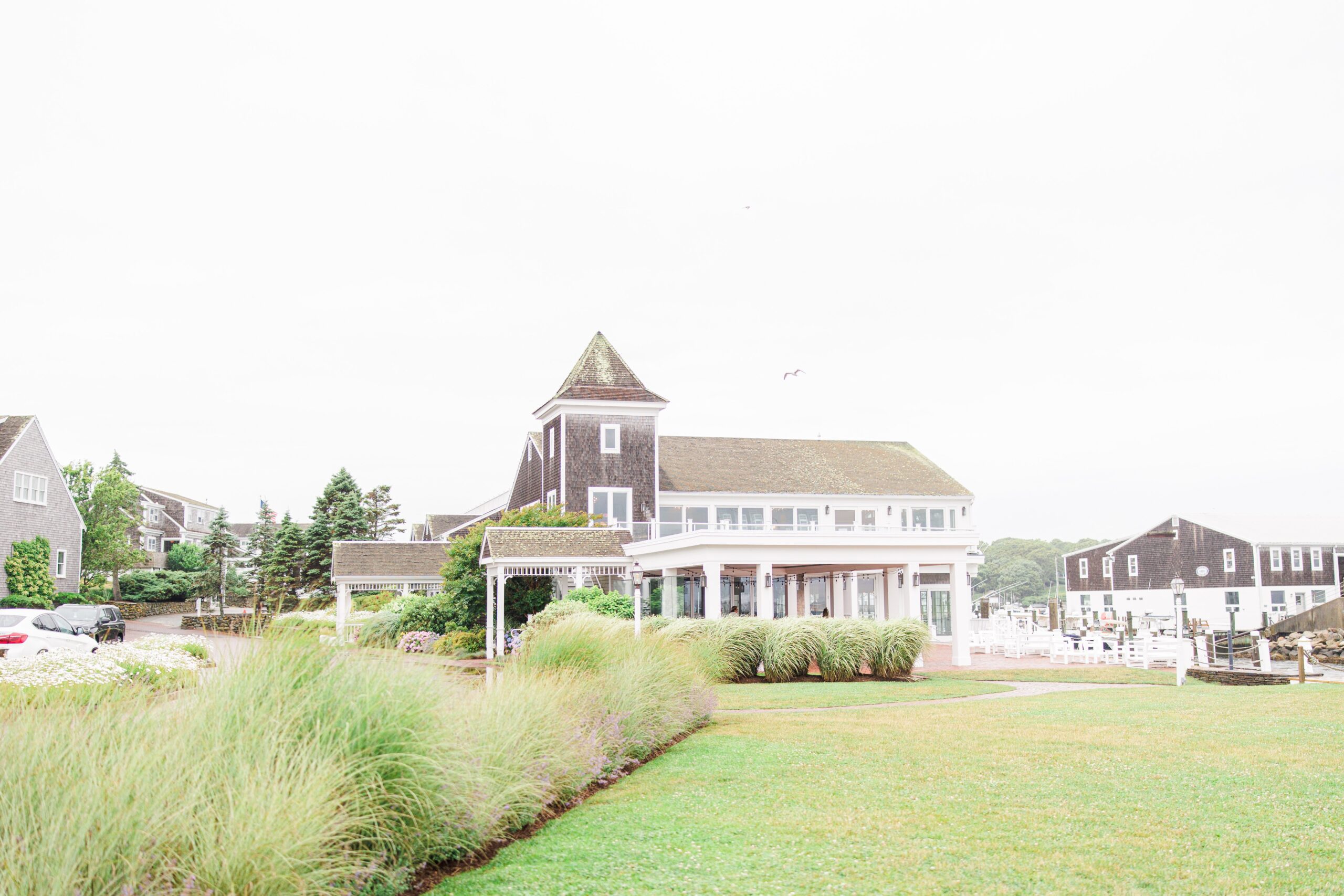 A charming Cape Cod inn at Wychmere Beach Club surrounded by manicured lawns and ornamental grasses. The building has classic New England architecture, with white trim and a pitched roof, set against a cloudy sky.