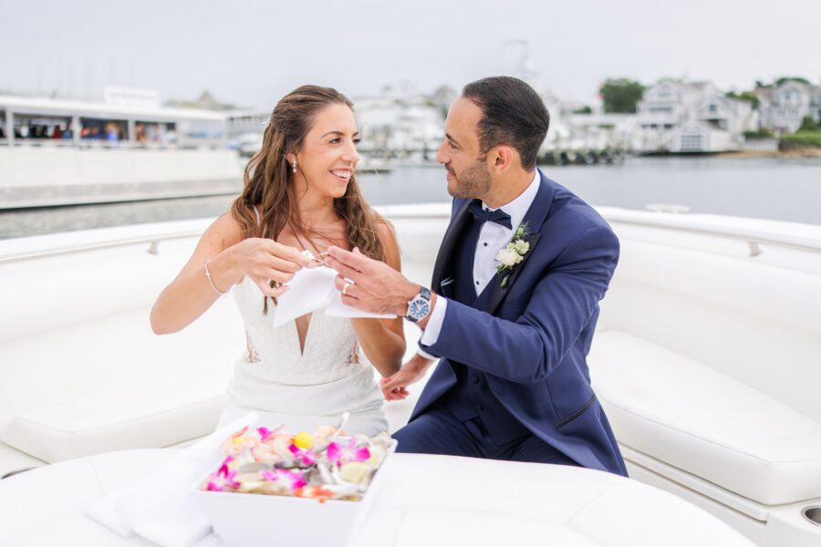 A bride and groom sit together on a white boat in Cape Cod. The groom, in a navy suit and bow tie, holds a small object while the bride, in a white lace dress, smiles at him. A bowl of colorful petals sits on the table in front of them. The backdrop features calm water and buildings, capturing a serene moment. Weddings on the Cape.