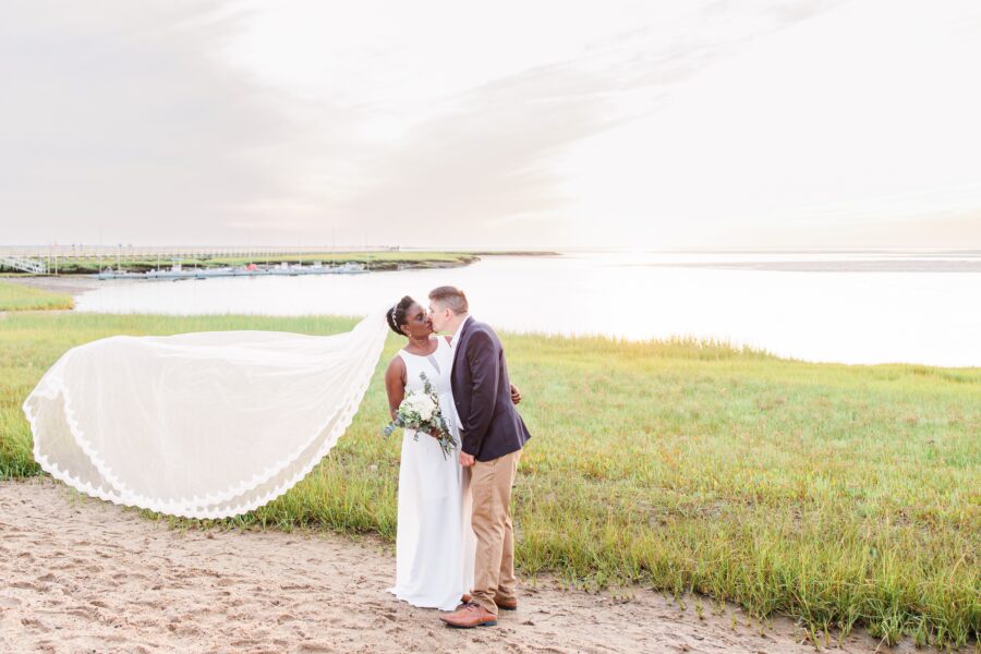 A bride and groom kiss on a grassy shoreline in Cape Cod at sunset. The bride’s veil billows gracefully behind her, and she holds a bouquet of white flowers. The groom wears a navy jacket and khaki pants. The glowing sky and water create a romantic scene. Weddings on the Cape.