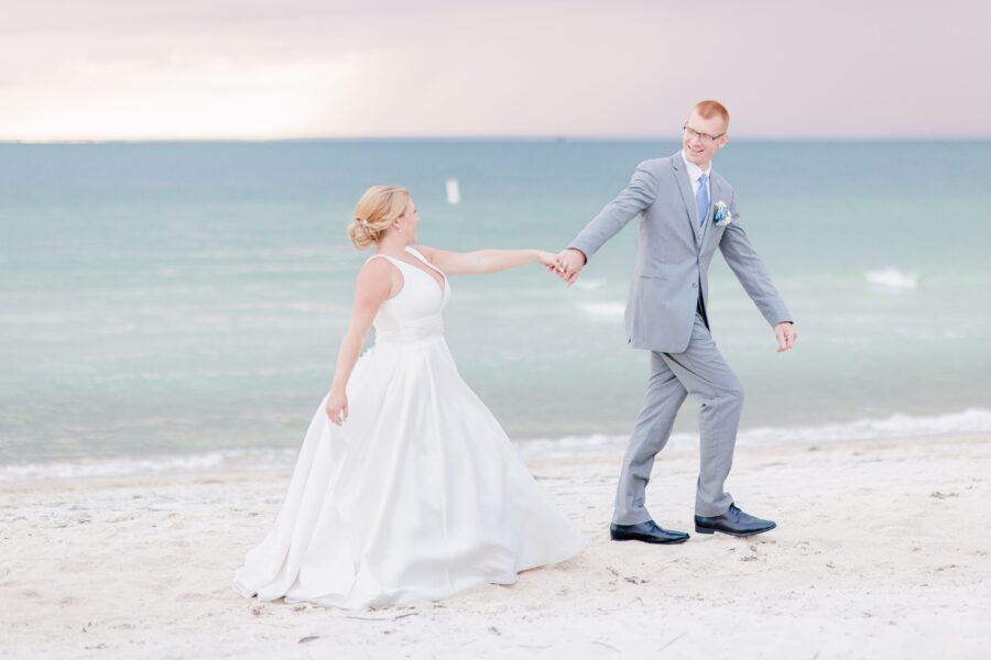 A bride and groom walk hand-in-hand on a sandy Cape Cod beach during their wedding. The bride wears a flowing white gown, and the groom is dressed in a light gray suit with a blue tie. The ocean stretches behind them under a soft, cloudy sky. Weddings on the Cape.