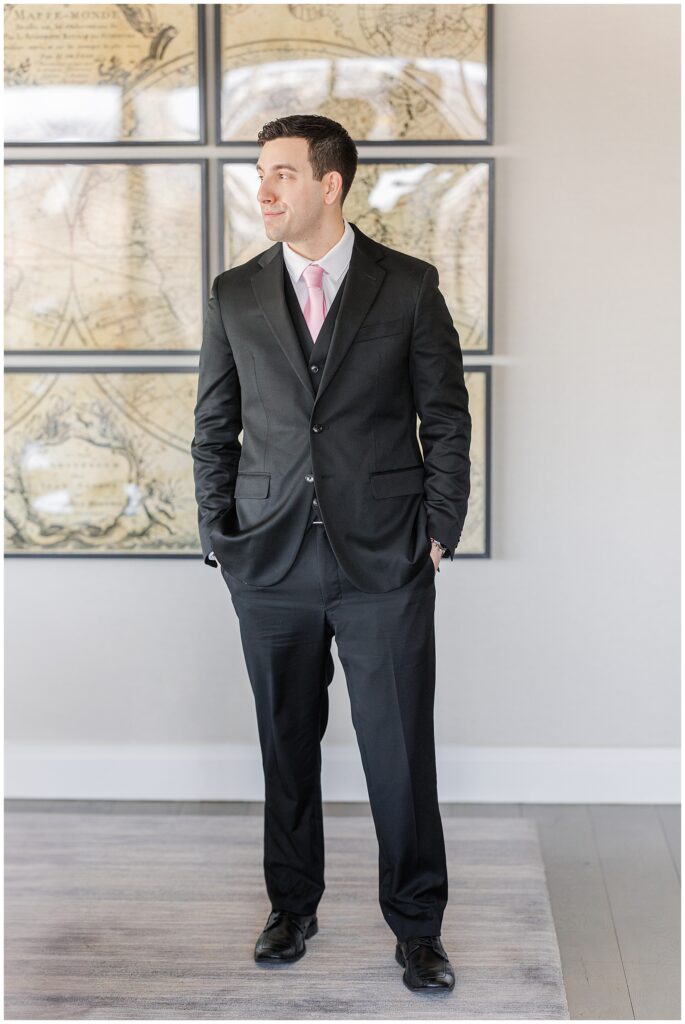 The groom, dressed in a black suit with a pink tie, stands in front of a vintage world map artwork in the presidential suite during his Ritz Carlton Boston wedding, gazing off to the side with a confident expression.