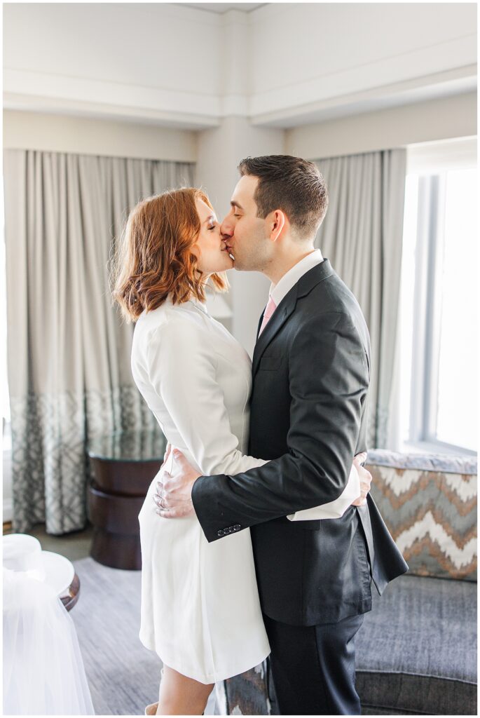 The newlyweds share a kiss in the presidential suite of the Ritz Carlton Boston, with natural light filtering through elegant curtains in the background.