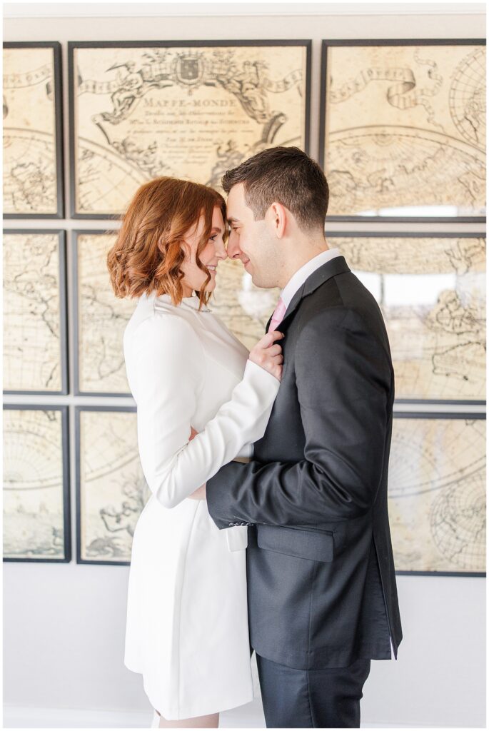 The couple stands close together, foreheads touching, as they share a tender moment during their Ritz Carlton Boston wedding. The groom wears a black suit and pink tie, while the bride wears a stylish white dress. A vintage world map artwork decorates the wall behind them.