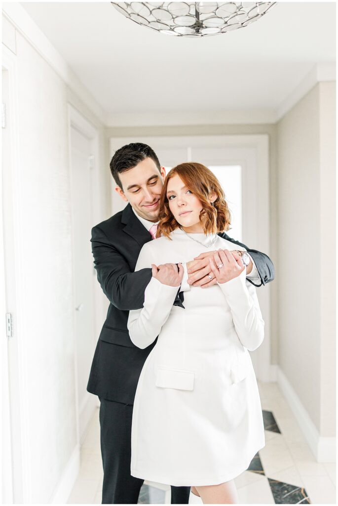 The groom stands behind the bride, wrapping his arms around her in a loving embrace during their Ritz Carlton Boston wedding. The bride gazes into the camera while the groom smiles softly against her shoulder.