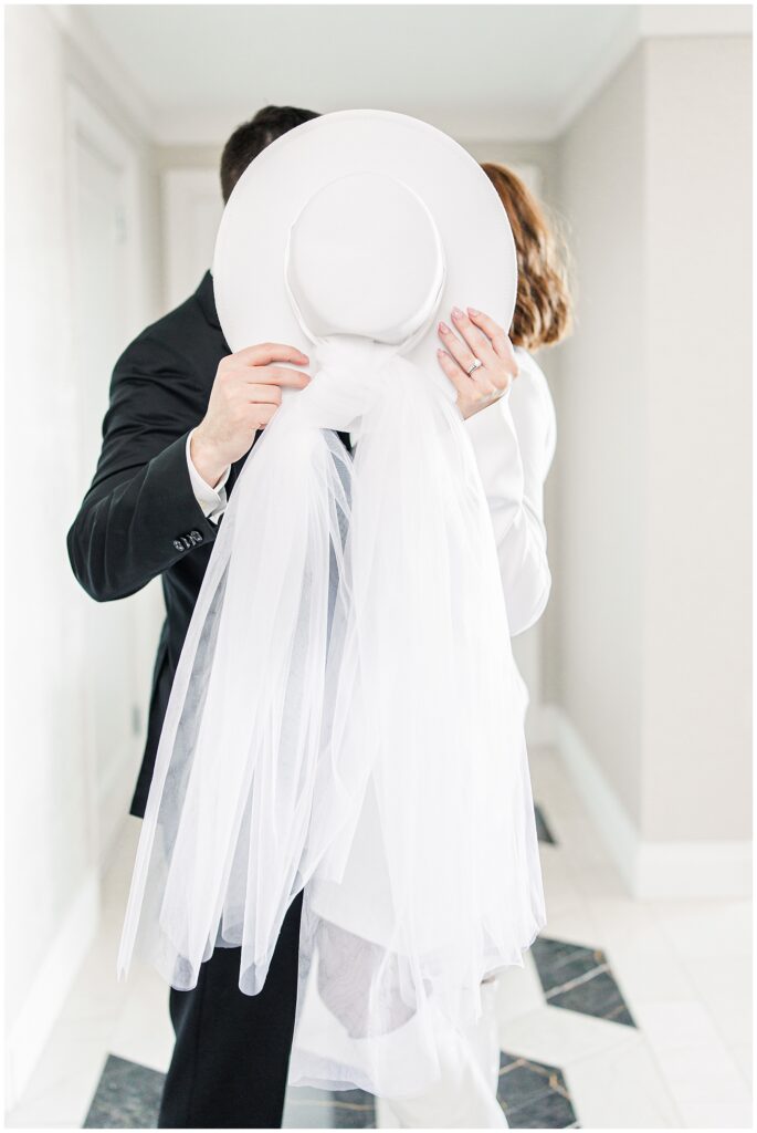 The groom holds a white bridal hat with a flowing veil in front of their faces as he and the bride share a private moment, their hands intertwined.