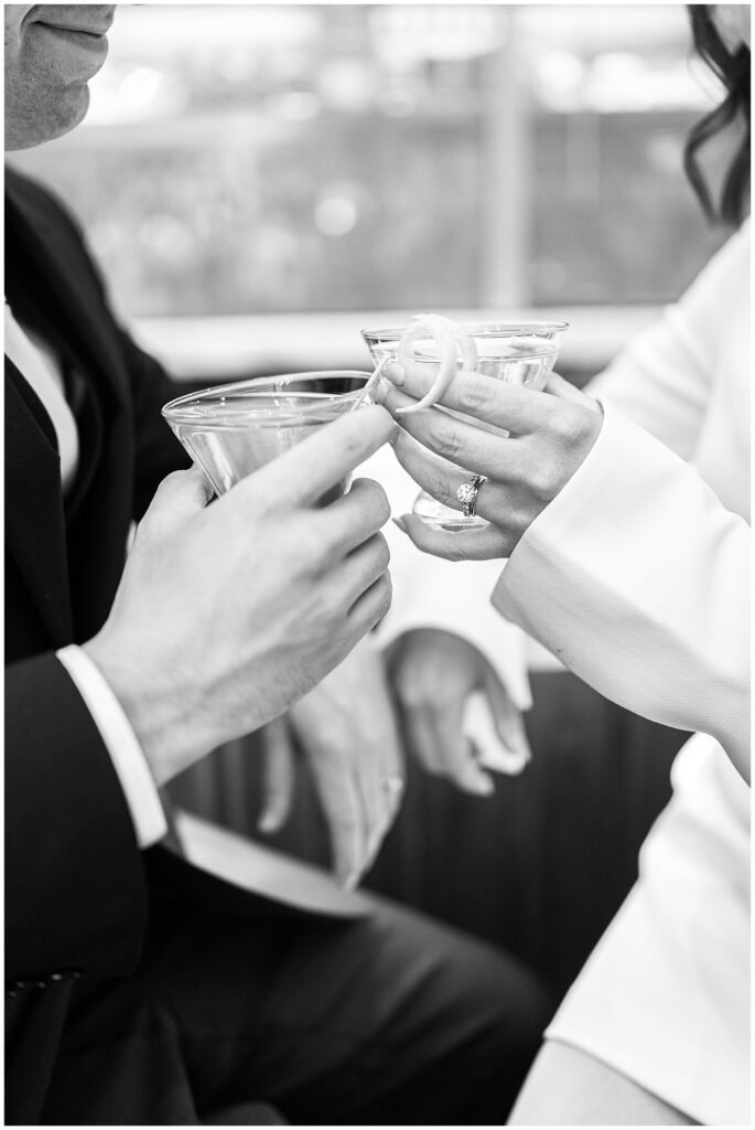 A close-up in black and white of the couple’s hands holding martini glasses with lemon twists. The bride’s engagement ring is prominently displayed as they toast during their Ritz Carlton Boston wedding