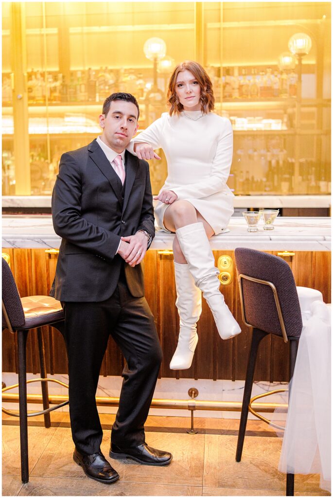 The couple poses together at the Avery Bar, with the groom standing confidently in front while the bride sits on the counter with her hand resting on his shoulder. Two martini glasses with garnishes sit on the marble counter.