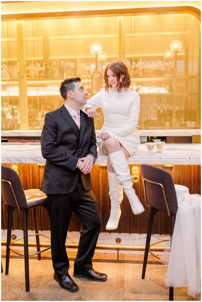 A bride sits on the marble bar counter at the Ritz Carlton Avery Bar, dressed in a chic white mini dress and knee-high boots, while the groom in a black suit with a pink tie leans on the bar, looking at her. The warm golden glow of the backlit bar adds to the ambiance.