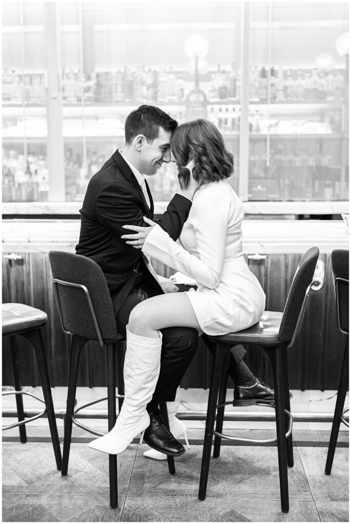 A black and white image of the bride and groom sitting close together at the bar, touching noses and smiling. The groom is in a black suit with a pink tie, while the bride wears a white mini dress and knee-high boots.