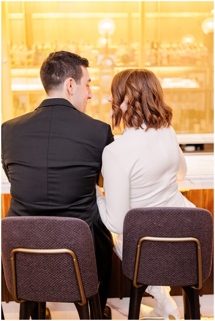 The couple sits side by side at the bar, facing away from the camera, engaged in conversation. The warm golden lighting highlights the bride’s white dress and the groom’s black suit during their Ritz Carlton Boston wedding.