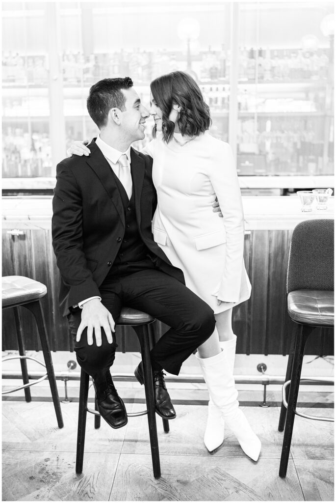 A black and white image of the couple sitting on barstools, leaning in with their foreheads touching during their Ritz Carlton Boston wedding. The bride’s knee-high boots and stylish mini dress contrast against the groom’s formal black suit.