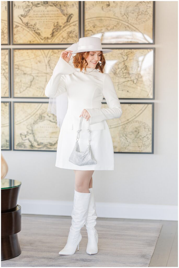 A bride in a modern white mini dress, white knee-high boots, and a white hat with a veil poses in the presidential suite. She holds a silver handbag and smiles while adjusting her hat. A vintage world map artwork is displayed behind her.