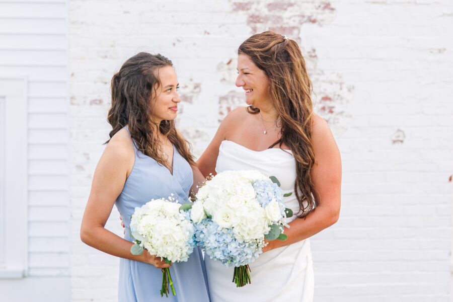 Two women smiling at each other while holding bouquets of white and blue flowers; one is in a white wedding dress, and the other is in a light blue bridesmaid dress, standing against a white brick wall.
