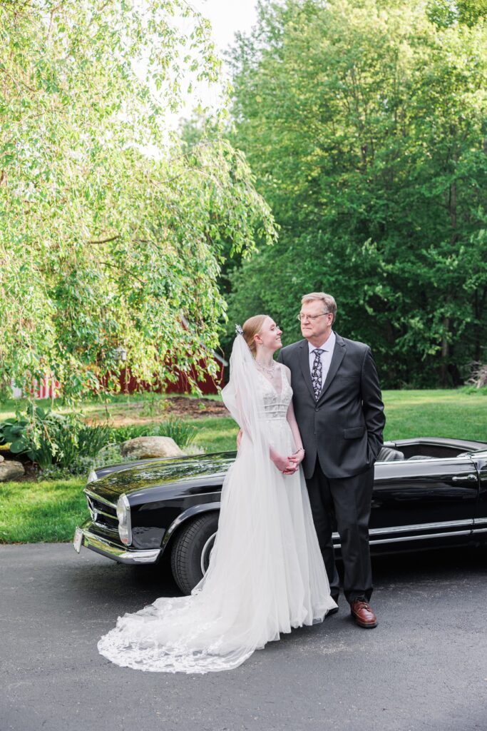 Bride in a white wedding gown and veil stands beside her father in a suit, posing in front of a vintage black convertible surrounded by trees—part of sentimental family wedding photos.