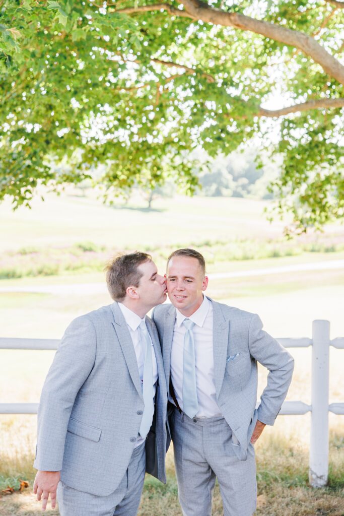 Two men in light gray suits standing outdoors; one kisses the other on the cheek under a tree, with a wooden fence and open field in the background.