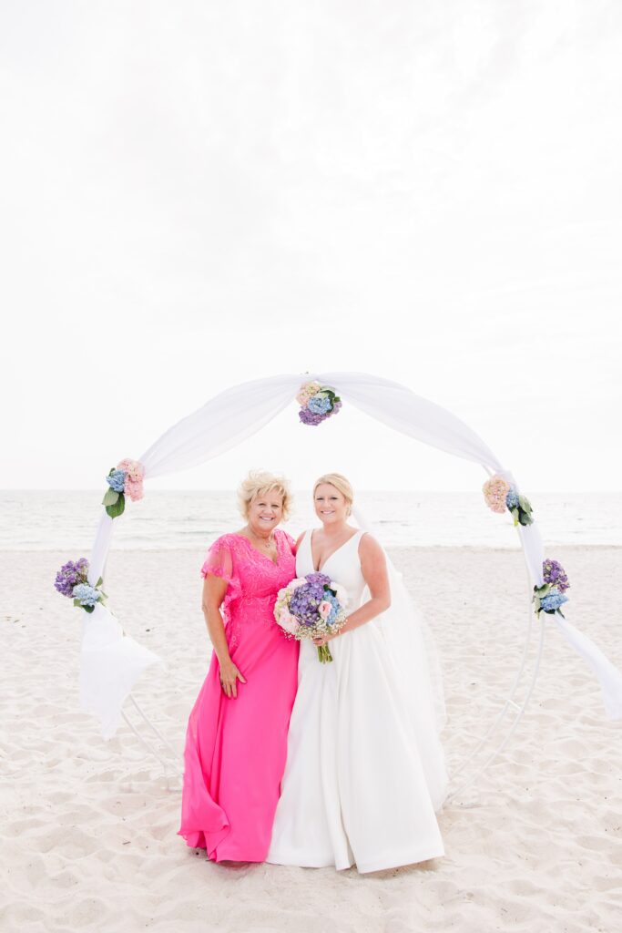 Bride and her mother in a bright pink dress standing on a beach under a floral arch, holding bouquets and smiling for relaxed family wedding photos.
