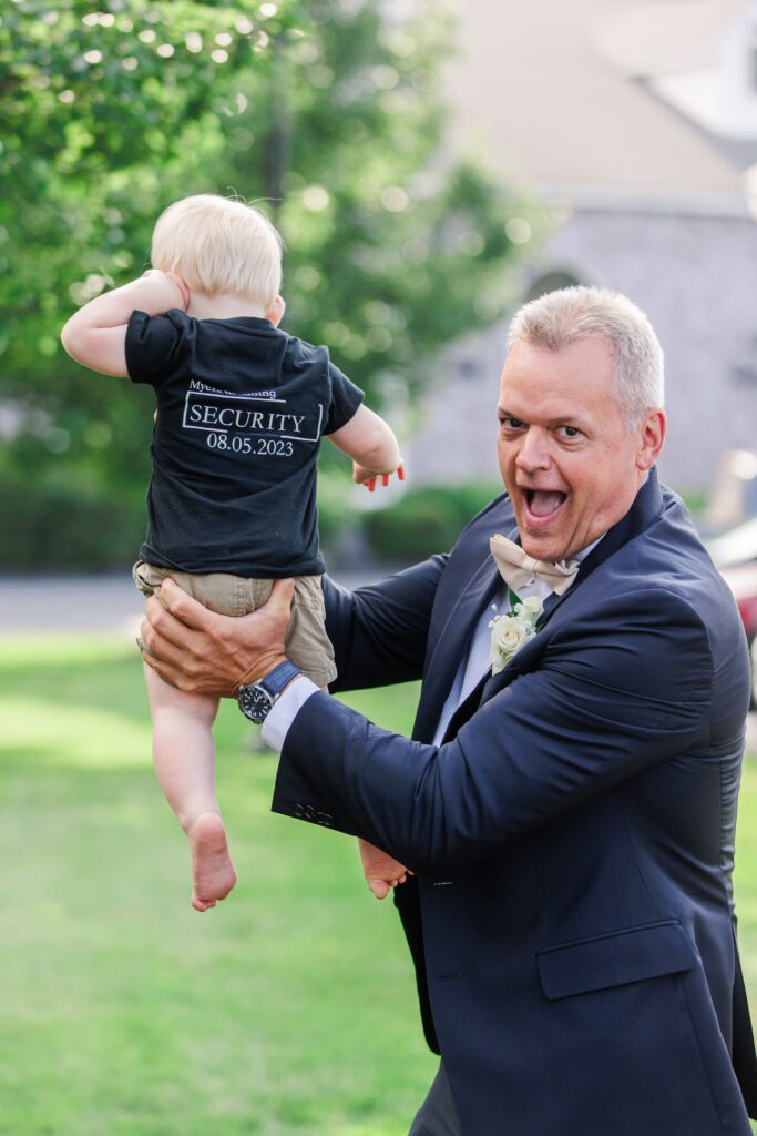 Man in a suit holding a small child wearing a shirt that reads “Security 08.05.2023,” both smiling and playful outdoors at a wedding event.