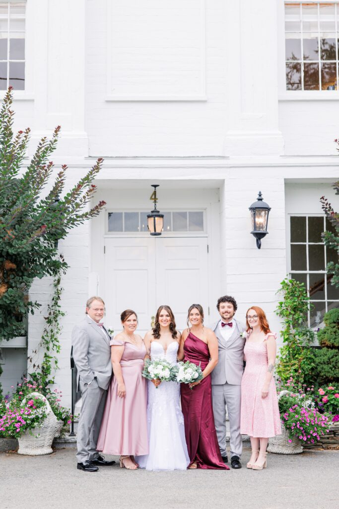 Bride in a white gown stands with five family members dressed in formal attire, posing in front of a white building adorned with flowers—part of elegant family wedding photos.