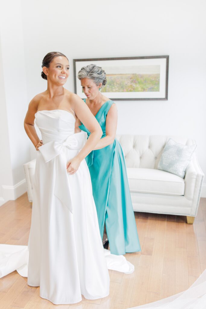 Bride smiling as an older woman in a teal dress helps fasten her wedding gown in a bright room with a white couch and framed artwork in the background.