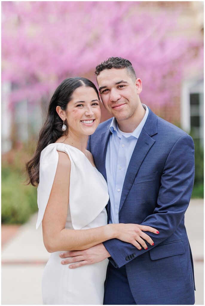 A close-up of the couple posing together, both smiling. The woman wears a white one-shoulder dress and the man wears a blue suit, with pink blossoms and a red-brick wall behind them.