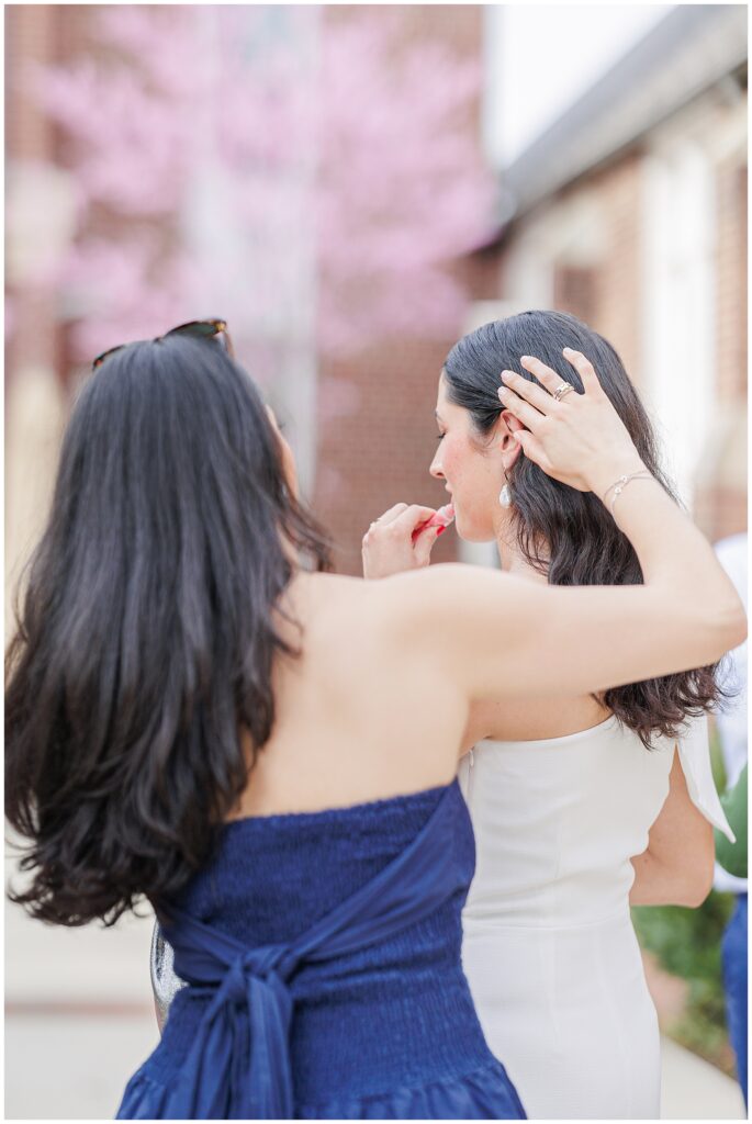 A woman in a navy strapless dress assists another woman in applying lipstick, adjusting her hair as they stand outdoors at the National Cathedral School, with pink-blossomed trees in the background.