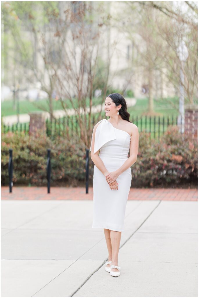 A woman in a fitted white dress stands smiling and looking to the side on a paved walkway, surrounded by trimmed shrubs, trees, and campus fencing at the National Cathedral School.