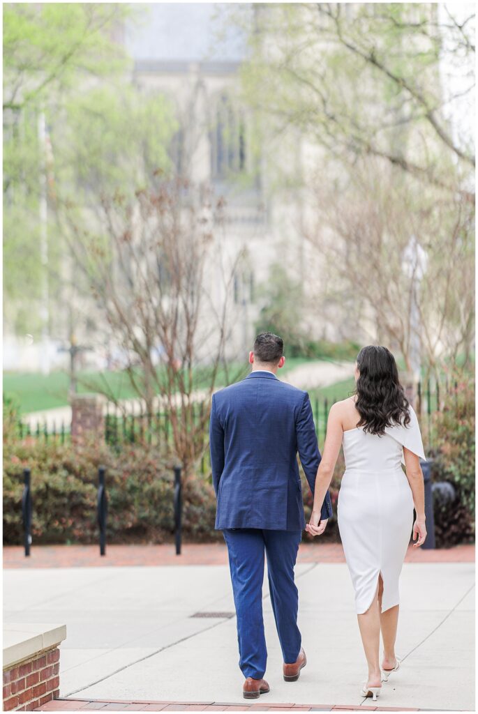 The couple walks away from the camera along a paved sidewalk, holding hands. They are surrounded by spring foliage and a blurred view of a large stone building in the distance.