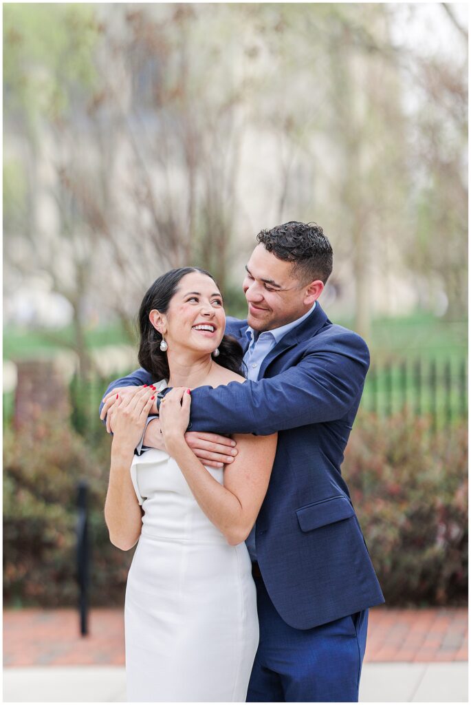 The man hugs the woman from behind as she smiles up at him. They are dressed formally and stand in front of foliage with the National Cathedral School campus in the background.