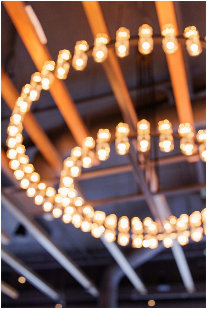 A close-up of two circular chandeliers with glowing bulbs, softly focused against a dark ceiling with exposed beams.
