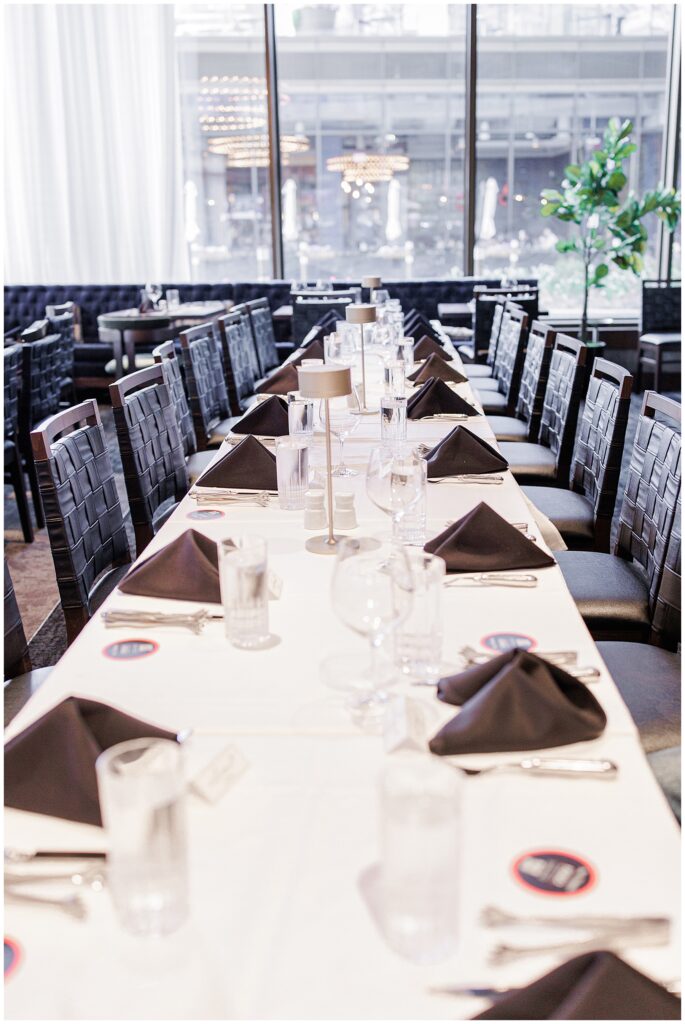 A long formal dining table inside Fogo de Chão at The Wharf, set with black napkins, clear glassware, and name cards, under large windows.