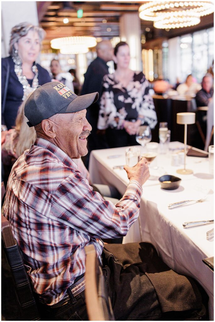 An older man in a plaid shirt and veteran cap sits at a dinner table smiling, surrounded by people at a warmly lit indoor event.