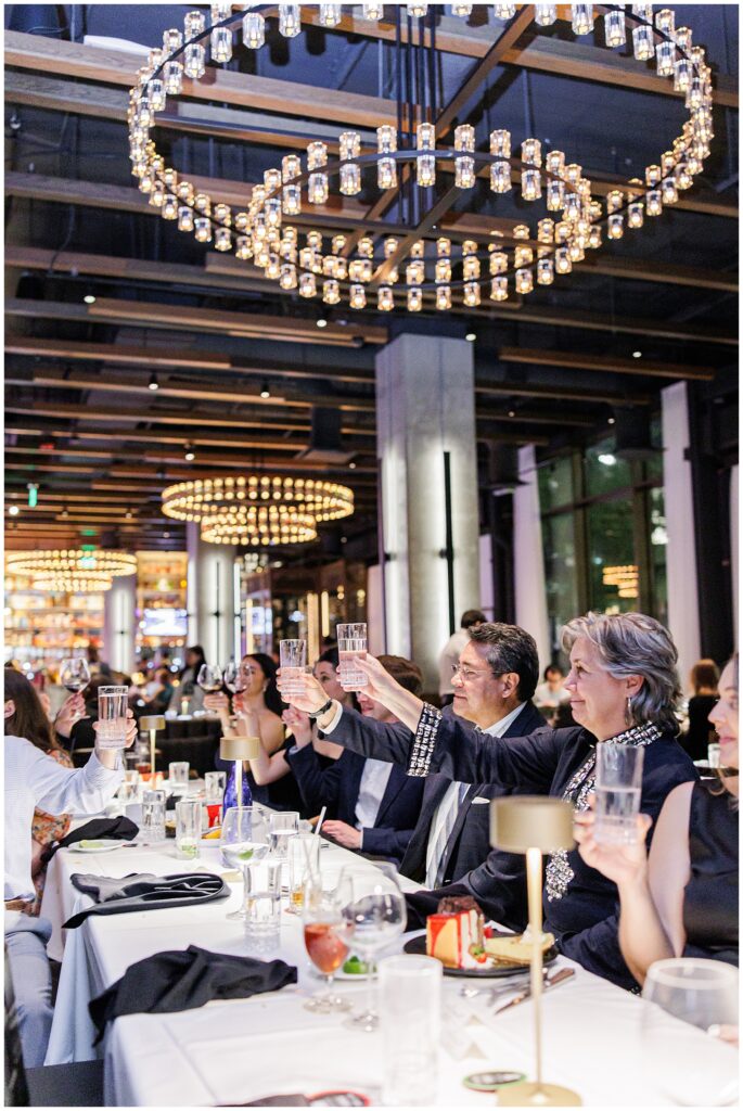 A group of guests sitting at a long table inside Fogo de Chão at The Wharf, raising glasses in a celebratory toast beneath ring-shaped chandeliers.