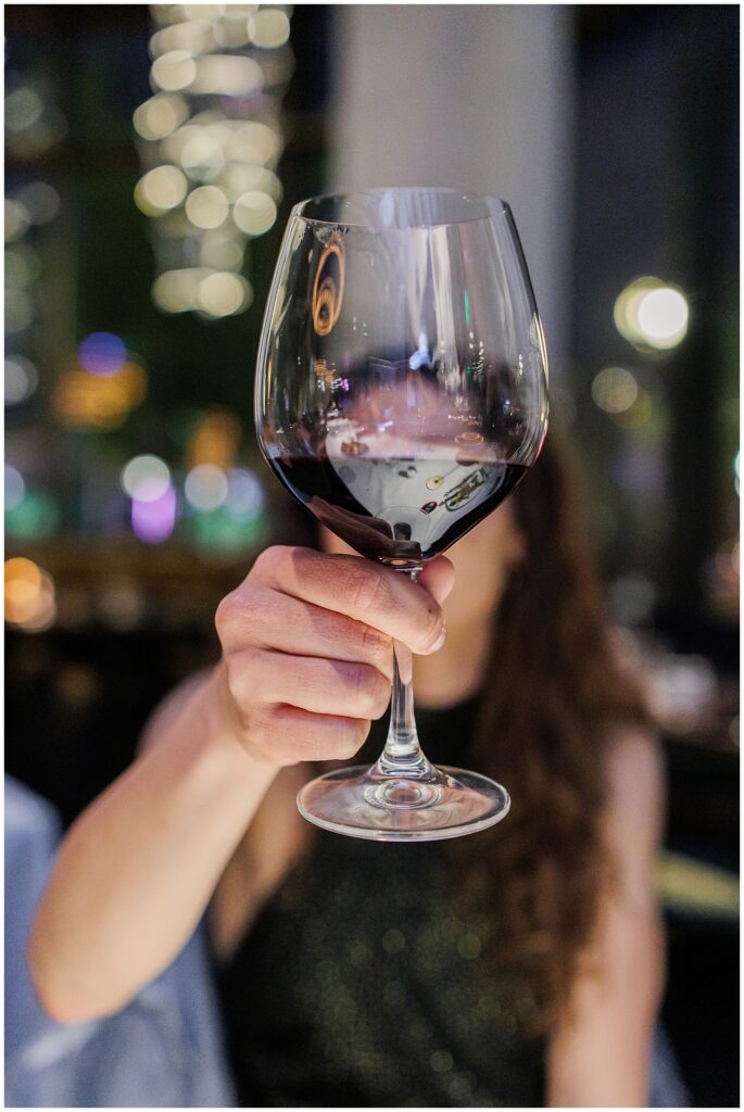 A close-up of a woman’s hand holding a red wine glass toward the camera, with a blurred background of lights and reflections.