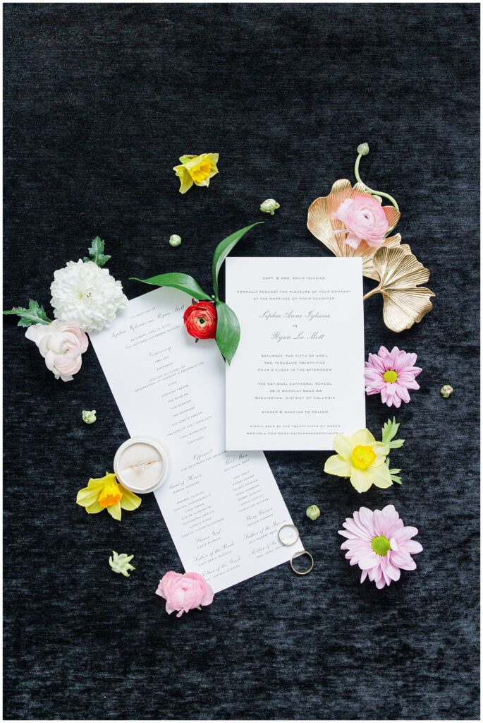 A styled flat lay of wedding invitations, ceremony programs, an engagement ring in a round box, and scattered colorful flowers on a dark fabric background, photographed by a Washington D.C. wedding photographer.
