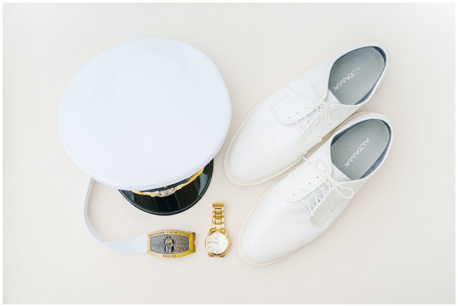 Flat lay of groom’s attire including a white military dress cap, white dress shoes, gold wristwatch, and a white belt with a “Cryptologic Warfare” insignia buckle.