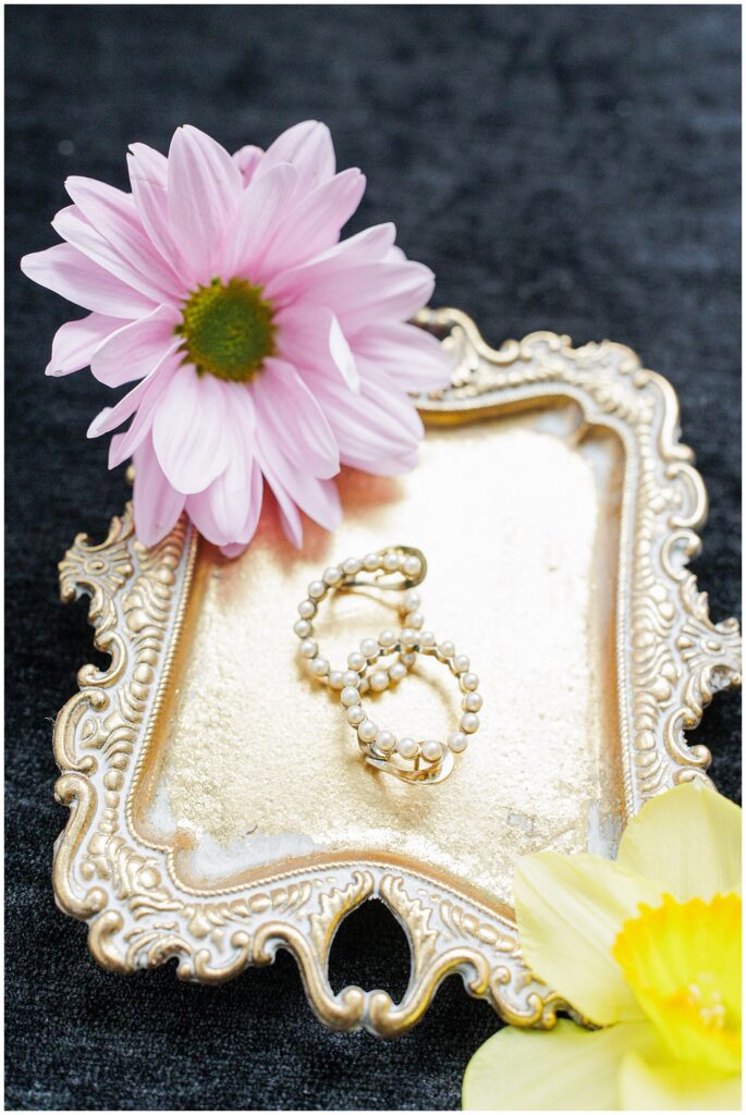 A pair of gold hoop earrings with pearls displayed on an ornate gold tray, with pink and yellow flowers on a dark fabric surface.