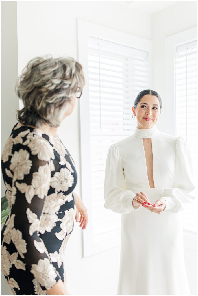 A bride in a long-sleeved white gown with a deep neckline stands smiling at an older woman in a floral black dress, in a naturally lit room with white blinds.