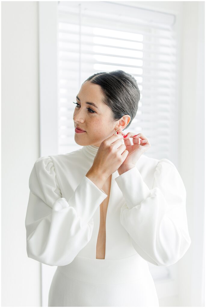 A bride with neatly styled hair and a white high-neck gown puts on pearl earrings near a bright window, photographed by a Washington D.C. wedding photographer.